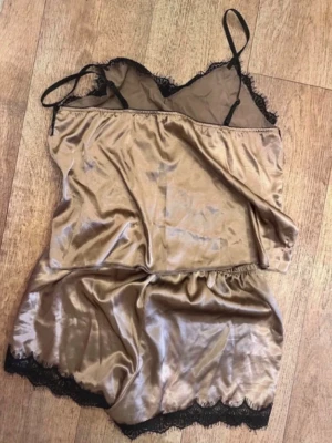 Sidenliknande pyjamasset med spets - Snyggt tvådelat pyjamasset i glansigt beige material med svarta spetsdetaljer. Setet består av ett linne med tunna axelband och V-ringning samt matchande shorts med elastisk midja. Perfekt för en lyxig känsla hemma.