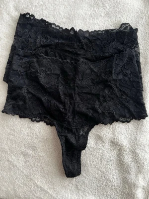 Svarta spetstrosor med hög midja - Ny pris 220kr. Snygga svarta trosor i mjuk spets med hög midja och breda sidor. Trosorna har blommigt spetsmönster och en bekväm passform. Perfekta för dig som gillar en trendig och feminin stil.