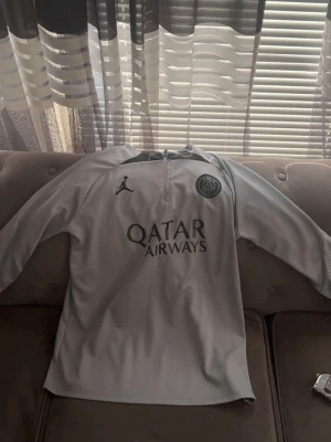 Grå PSG träningströja Jordan - Snygg grå långärmad träningströja från PSG och Jordan med halv dragkedja framtill. Tröjan har PSG-logga och Jordan-logga på bröstet samt Qatar Airways-tryck. Tillverkad i ett lätt och funktionellt material som passar perfekt till träning.