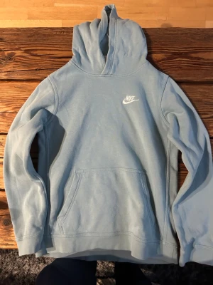 Ljusblå hoodie från Nike - Snygg ljusblå hoodie från Nike med klassisk logga på bröstet. Hoodien har huva och en stor magficka framtill. Tillverkad i mjukt material som känns skönt mot huden. Perfekt för en avslappnad och sportig stil. Köpt från JD sports. 