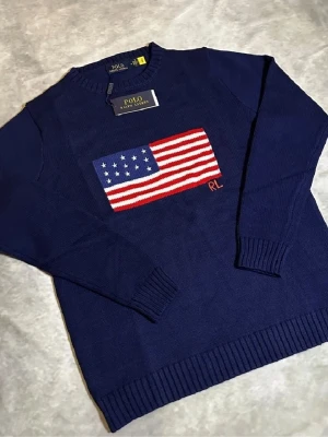 Mörkblå stickad tröja Polo Ralph Lauren - Mörkblå stickad tröja från Polo Ralph Lauren med amerikansk flagga i rött, vitt och blått på bröstet samt RL-broderi. Tröjan har rund halsringning och ribbade muddar vid ärmslut och nederkant. Perfekt för dig som gillar klassisk och stilren design.