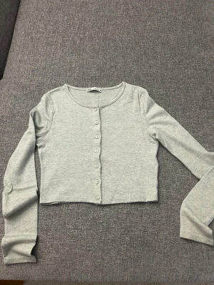 Ljusgrå cropped kofta från Zara - Ljusgrå cropped kofta från Zara med långa ärmar och rund halsringning. Koftan har knappar framtill och är gjord i ett mjukt, stretchigt material. Perfekt att styla över ett linne eller t-shirt.