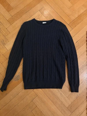 Mörkblå kabelstickad tröja från John Henric - Mörkblå kabelstickad tröja från John Henric med rund halsringning och långa ärmar. Tröjan har ribbade muddar vid ärmslut och nederkant, vilket ger en klassisk och stilren look. Perfekt för dig som gillar enkel men snygg design.