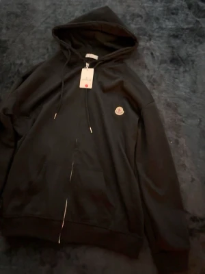 Svart hoodie från MC - Svart hoodie från MC med dragkedja framtill, justerbar huva med snören och klassisk M-logga på bröstet. Tillverkad i mjukt material som känns skönt mot huden. Perfekt för en avslappnad och stilren look.