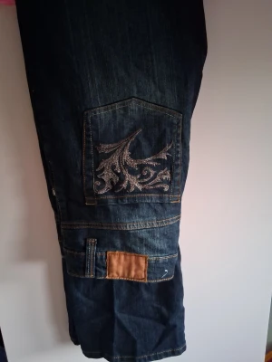 Blå jeans med broderad ficka - Snygga blå jeans med unik broderad ficka på benet och kontrastsömmar. Jeansen har en extra detalj med en brun patch och praktiska fickor. Perfekta för dig som vill sticka ut med coola detaljer.