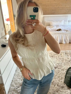 Krämvit blus med smock och volang - Supersöt krämvit blus med smockdetaljer och volang nertill. Ärmlös modell med rund halsringning och lätt rynkat tyg som ger en romantisk känsla. Perfekt att styla med jeans eller kjol för en fräsch look.