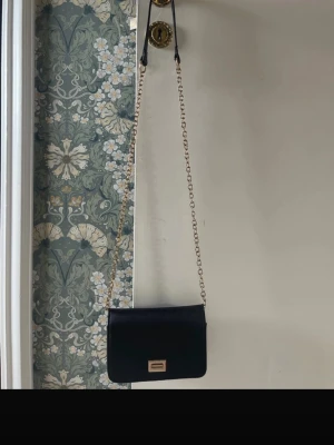 Primadonna axelremsväska  - Superfin svart axelremsväska med guldkedja från Primadonna, nästan helt nytt skick  Man stänger den med ett guldspänne se bild 3-4  Litet fack med dragkedja inuti se bild 5  Säljer då den inte kommer till användning 💕 Hör av dig vid frågor ❣️❣️❣️