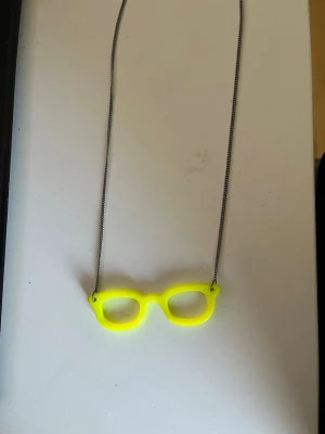 Halsband med neon-gula glasögon - Unikt halsband med en kedja i metall och ett hänge i form av neon-gula glasögon. Hänget är gjort av plast och har en lekfull, ungdomlig design som sticker ut. Perfekt för dig som gillar färgglada accessoarer och vill addera en cool detalj till din outfit.