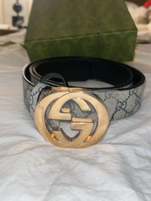 Gucci GG Supreme bälte med guldspänne - Snyggt Gucci GG Supreme bälte med klassiskt guldigt GG-spänne. Bältet har det ikoniska GG-mönstret i grått och beige och är tillverkat i skinn med canvasdetaljer. Perfekt accessoar för att lyfta vilken outfit som helst.