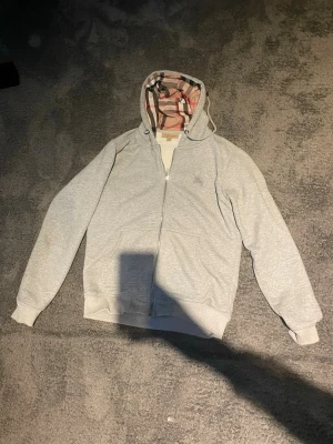 Grå zip hoodie  - Snygg grå zip hoodie med klassiskt rutigt foder i huvan och diskret broderad logga på bröstet. Hoodien har dragsko i huvan, två fickor fram och ribbade muddar. Perfekt för en chill och stilren look.