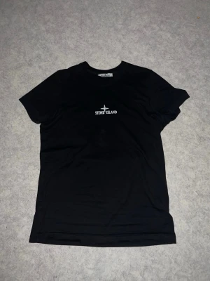 Stone Island t-shirt med tryck - Cool svart t-shirt från Stone Island med vit logotyp på bröstet och ett stort grafiskt tryck i brunt, gult och vitt på ryggen. Klassisk passform och rund hals. Perfekt för dig som gillar streetwear och snygga detaljer.