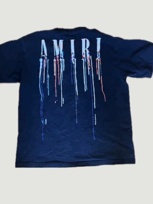 Mörkblå AMIRI t-shirt med tryck - Snygg mörkblå t-shirt från AMIRI med färgstarkt drip-tryck både fram och bak. Stor AMIRI-logga på ryggen med färgstänk i blått, vitt och rött. Klassisk passform och rund hals. Perfekt för dig som gillar streetwear och vill sticka ut.