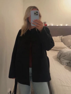 Svart kappa med knytskärp - Stilren svart kappa med knytskärp i midjan och rak passform. Kappan har rund halsringning och stora fickor framtill. Perfekt för dig som vill ha en enkel men trendig look. Passar till jeans och sneakers för en avslappnad stil.