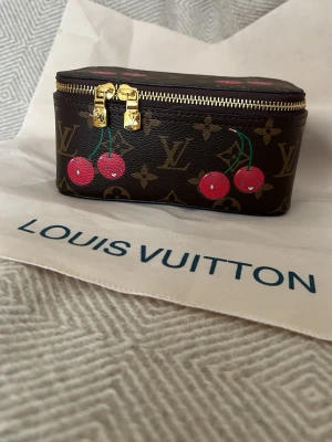  necessär med körsbär - Säljer en brun necessär  med klassiskt monogrammönster och söta körsbärsdetaljer i rött och grönt. Väskan har guldfärgade dragkedjor och en kompakt, rektangulär form. Perfekt för smink eller småprylar.