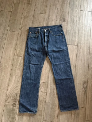 Levi's 501 blå raka jeans - Klassiska blå Levi's 501 jeans med rak passform och fem fickor. Jeansen har knappgylf och är gjorda i slitstark denim. Perfekta för en avslappnad och tidlös stil. Ikonisk läderpatch bak i midjan.
