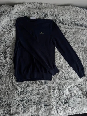 Mörkblå tröja från Lacoste - Snygg mörkblå tröja från Lacoste med klassisk rund halsringning och den ikoniska krokodilloggan broderad på bröstet. Tröjan har långa ärmar och ribbade muddar vid ärmslut och nederkant. Perfekt för en clean och stilren look.