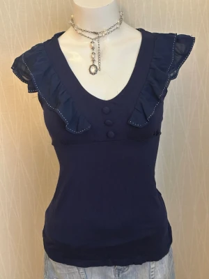 Mörkblå topp med volang och knappar - Supersnygg mörkblå topp med v-ringning, volangdetaljer längs axlarna och tre dekorativa knappar framtill. Ärmlös modell med lätt transparent volangkant och figurnära passform. Perfekt att styla med jeans eller kjol.