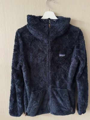 Patagonia fleecejacka mörkblå L herr – mycket fint skick - Patagonia fleecejacka / hoodie i mörkblå / navy, storlek L herr. Mycket fint begagnat skick. Mjuk och varm modell med huva, hel dragkedja framtill och sidofickor med zip. Perfekt till vardag, layering och outdoor. Klassisk Patagonia-piece i lättmatchad färg.