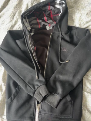 Svart hoodie med rutigt foder - Säljer en svart hoodie från Burberry med dragkedja och klassiskt rutigt foder i huvan. Hoodien har fickor framtill, ribbade muddar och dragsko i huvan. Perfekt för dig som gillar stilrena plagg med en twist. Materialet känns mjukt och bekvämt.