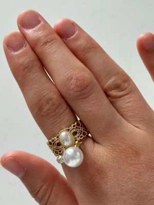 Guldfärgad ring med pärlor och strass - Unik guldfärgad ring med ett öppet, flätat band och dekorativa detaljer. Ringen har två vita pärlor i olika storlekar och en liten gnistrande strass. Perfekt för dig som gillar statement-smycken och vill sticka ut lite extra.