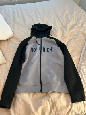 Svart och grå hoodie från Hoodrich - Säljer en snygg hoodie från Hoodrich i svart och grått med dragkedja framtill och stor huva med snörning. Hoodien har tryckt logga på bröstet och två fickor fram. Perfekt för en chill och street-inspirerad look.