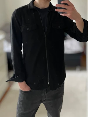 Svart overshirt från Buratti | L - Stilren svart overshirt från Buratti med dragkedja framtill och två bröstfickor med knapp. Overshirten har klassisk krage och är tillverkad i ett mjukt tyg som passar perfekt till lager-på-lager. Enkel att matcha med olika outfits.