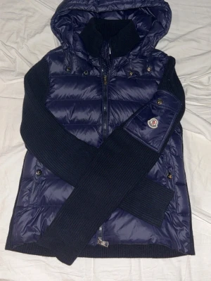 Mörkblå Moncler cardiganjacka med luva  - Snygg mörkblå cardiganjacka från Moncler med luva. Ärmar och rygg är stickade i mjukt material, medan framsidan är glansig och vadderad. Dragkedja framtill. Jackan är storlek S. Jag säljer jackan för jag tyckte den va för kort på mig. Jackan är i helt nytt skick och har inga tecken på användning. Förutom att ena sidan på den lilla tyg biten innne i jackan  lossnade när jag skulle dra bort prislapp inne i jackan. Nfc tag funkar som den ska.Pris går att diskutera!