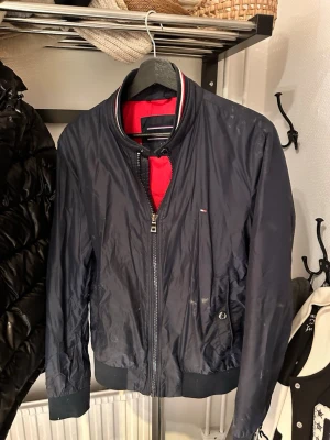 Marinblå vindjacka från Tommy Hilfiger - Snygg marinblå vindjacka från Tommy Hilfiger med röd innerfoder och klassiska Hilfiger-detaljer på kragen. Jackan har dragkedja framtill, två sidofickor med knapp och ribbade muddar vid ärmslut och nederkant. Perfekt för blåsiga dagar.