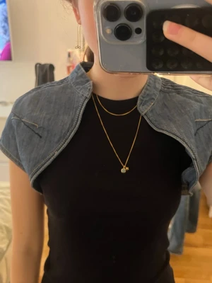 Kort jeansväst  - Säljer en kort, bolero-inspirerad jeansväst. Västen är i blå denim med markerade sömmar och har en öppen front utan knappar. Perfekt för att ge din outfit en cool och trendig touch.