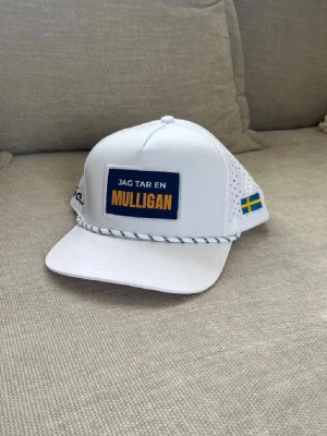 Vit keps Golfio.se Mulligan - Vit keps med rak skärm och broderad text 'Jag tar en Mulligan' framtill. Sidorna har perforerade hål för ventilation, svensk flagga på sidan och Golfio.se-brodyr. Dekorativt snöre framtill. Perfekt för golfbanan eller som cool accessoar.