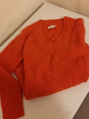 Orange stickad tröja från Kaffe - Mysig orange stickad tröja från Kaffe med djup v-ringning och långa ärmar. Tröjan har en fluffig, mjuk struktur och ribbstickade detaljer vid halsringningen. Perfekt för lager på lager och höstiga outfits.
