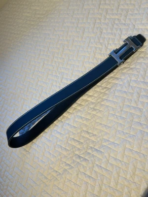 Hermesss blått skärp med H-spänne - Snyggt blått skärp från Hermesss med ikoniskt H-format metallspänne. Skärpet är gjort i skinn och har vita kontrastsömmar längs kanterna. Perfekt accessoar för att lyfta din outfit med en lyxig touch.