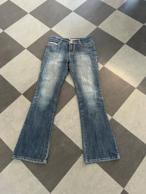Blå bootcut jeans med slitningar - Säljer ett par blå bootcut jeans med snygga kontrastsömmar. Jeansen har klassisk femficksdesign och lätt utsvängda ben. Perfekta för dig som gillar en retro och avslappnad stil. Materialet är denim i bomull.