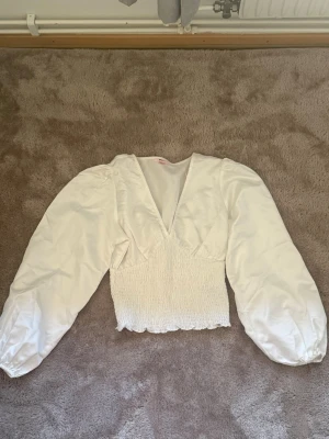 Vit blus med puffärm från Gina Tricot - Supersnygg vit blus från Gina Tricot med puffiga långärmar och smockad midja. Djup v-ringning och lätt glansigt material ger en trendig look. Har använt 1 gång