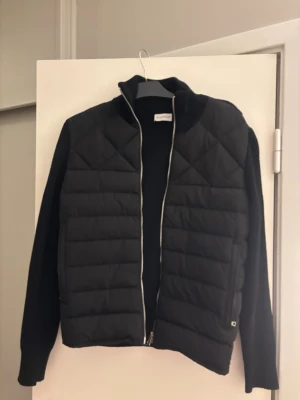 Svart pufferjacka från Moncler - Snygg svart pufferjacka från Moncler med quiltad front och stickade ärmar. Jackan har dragkedja framtill och en diskret Moncler-logga på ärmen. Perfekt för dig som vill ha en stilren och trendig look.