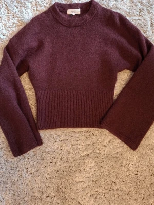 Gina tricot - Säljer en croppad stickad tröja i plommonlila färg med bred ribbstickad midja och vida ärmar. Tröjan har rund halsringning och är perfekt för lager på lager. Superskön och trendig till jeans eller kjol.