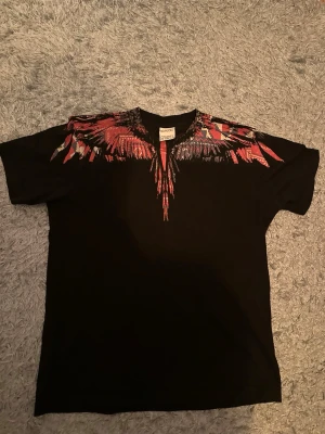 Svart t-shirt från Marcelo Burlon - Svart t-shirt från Marcelo Burlon med ikoniskt vingmönster i rött, rosa och vitt över axlar och halsringning. Klassisk passform och rund hals. Perfekt statement-plagg för dig som vill sticka ut.