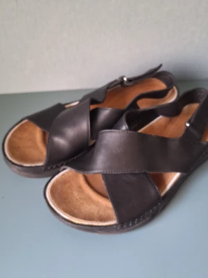 Clarks Trigenic svarta sandaler med korsade remmar - Clarks Trigenic - snygga svarta sandaler med breda korsade remmar i läder och justerbar hälrem med spänne. Innersulan är mjuk och beige, vilket ger en skön känsla för foten. Yttersulan är svart och robust, perfekt för sommardagar.