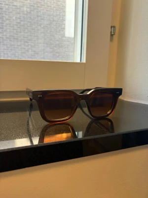 Bruna solglasögon från CHIMI - Snygga solglasögon från CHIMI med fyrkantig form och mörkbruna bågar. Glasen är tonade i brunt och bågarna har en stilren design med diskreta silvriga detaljer vid gångjärnen. Perfekta för att höja din look under soliga dagar.