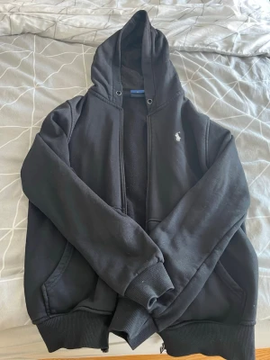 Svart hoodie från Polo Ralph Lauren - Svart hoodie från Polo Ralph Lauren med dragkedja, huva och fickor på sidorna. Klassisk vit logga broderad på bröstet. Tillverkad i mjukt bomullsmaterial med ribbade muddar vid ärmslut och nederkant. Perfekt för en avslappnad och stilren look.