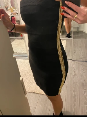 Svart axelbandslös kort klänning - Helt ny Snygg svart axelbandslös klänning med guldiga detaljer längs sidan. Tight passform och kort modell som framhäver figuren. Perfekt för dig som vill sticka ut med en elegant men edgy look.
