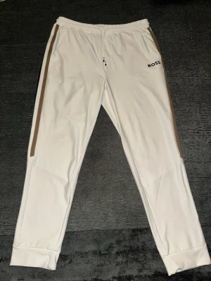 Boss Matteo Berrettini - Säljer ett par vita mjukisbyxor från BOSS med beige och svarta sidopaneler. Byxorna har elastisk midja, smal passform och diskret BOSS-logga på vänster sida. Perfekta för en chill och stilren look. Materialet känns mjukt och följsamt.