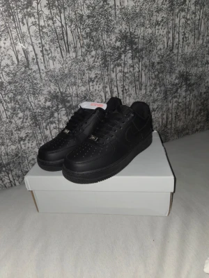 Helt nya nike air force 1 svarta - Säljer ett par klassiska Nike Air Force 1 sneakers i helsvart läder. Skorna har perforerad tå, svarta skosnören och metallbricka med 'AF1' på snörningen. Swoosh-logga på sidan och bekväm sula. Perfekta för dig som gillar stilrena och tidlösa sneakers.