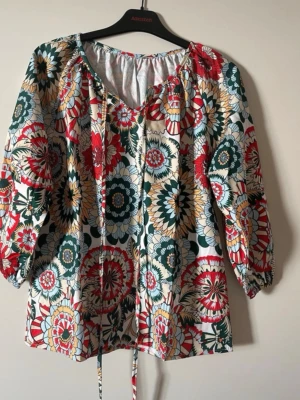 Flerfärgad blommig blus med knytband - Blus/ topp i härligt retromönster och färger. 3/4 arm med resår Knytband vid halsringningen. St M Längd från axel: 64 cm Bredd under armhål: 50 cm  100% polyester  Lite blank i tyget.  Oanvänd