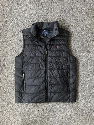 Svart dunväst från Polo Ralph Lauren - Snygg svart dunväst från Polo Ralph Lauren med horisontella sömmar och dragkedja framtill. Västen har två fickor med dragkedja och en liten röd logga på bröstet. Perfekt för lager-på-lager och håller dig varm under kyliga dagar. Storleken är L ungdom motsvarar Xs