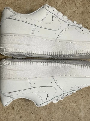 Nike Air Force 1 vita låga sneakers - Klassiska Nike Air Force 1 sneakers i helvitt läder med ikonisk swoosh på sidan. Lågt skaft, tjock sula och perforerade detaljer vid tån för extra ventilation. Perfekta för en clean och stilren look.