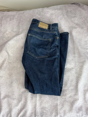 Mörkblå jeans från 157 - Snygga mörkblå jeans från 157 med klassisk femficksdesign och raka ben. Jeansen har en beige patch bak i midjan och är tillverkade i slitstark denim. Perfekta för en avslappnad och trendig look.