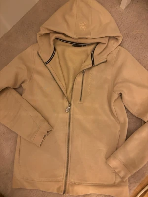 Beige hoodie från Sail Racing - Snygg beige hoodie från Sail Racing med dragkedja hela vägen, broderad logga på bröstet och huva med diskret emblem. Tillverkad i mjukt material, perfekt för chill dagar. Svart detalj på insidan av kragen ger extra stilpoäng.