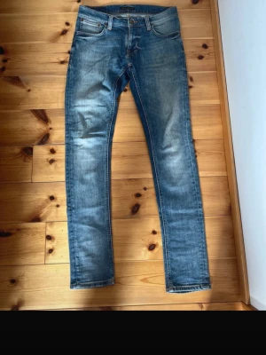 Blå slim fit jeans från Nudie Jeans, modell : thight terry - Säljer ett par blå slim fit jeans från Nudie Jeans med klassisk femficksmodell och snygga slitningar framtill. Jeansen har normal midja, dragkedjegylf och är tillverkade i mjuk denim. Perfekta för en avslappnad och trendig look.