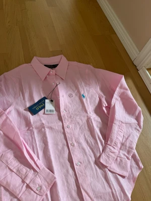 Rosa skjorta från Polo Ralph Lauren - Säljer en helt ny klassisk rosa skjorta från Polo Ralph Lauren med lapparna på! Den ikoniska loggan broderad på bröstet. Skjortan har lång ärm, knappar framtill och är gjord i material som känns som linne men är ej säker! Perfekt inför sommaren för dig som gillar stilrena och tidlösa plagg. Skickas självklart i originalförpackning!
